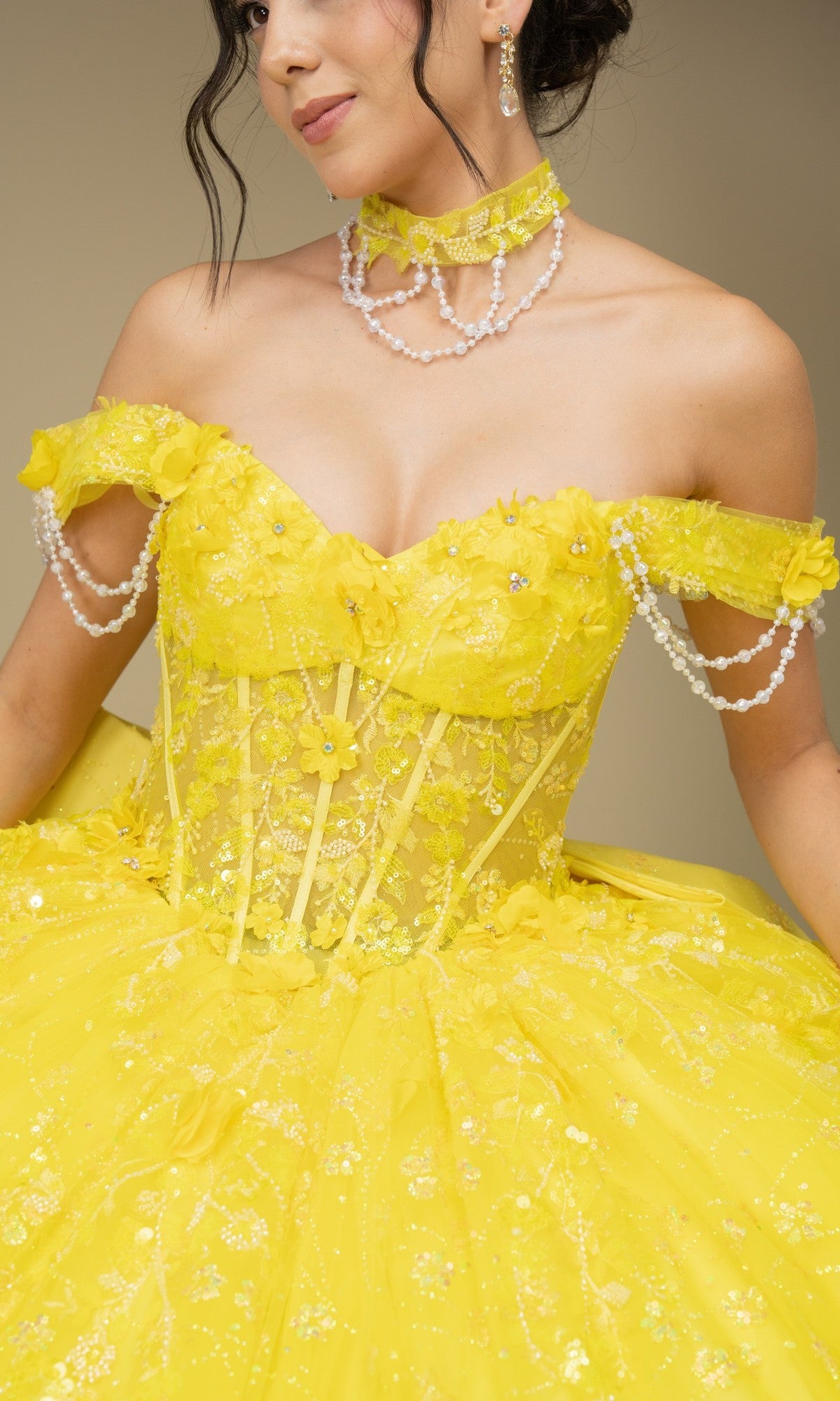 A woman wears the Quinceanera Dress 8415JQ by Cinderella Couture—an ornate, off the shoulder yellow gown with floral details, lace, pearl accents, and matching pearl jewelry for a fairytale-inspired look.